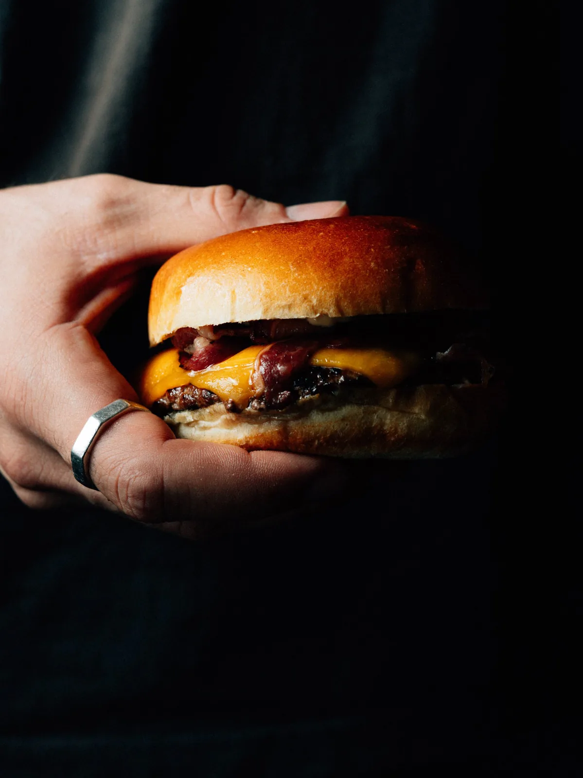 Smash burger bacon cheddar tenu en main sur fond noir, Brute Burger à Cavalaire-sur-Mer, photo Hugz Visuals
